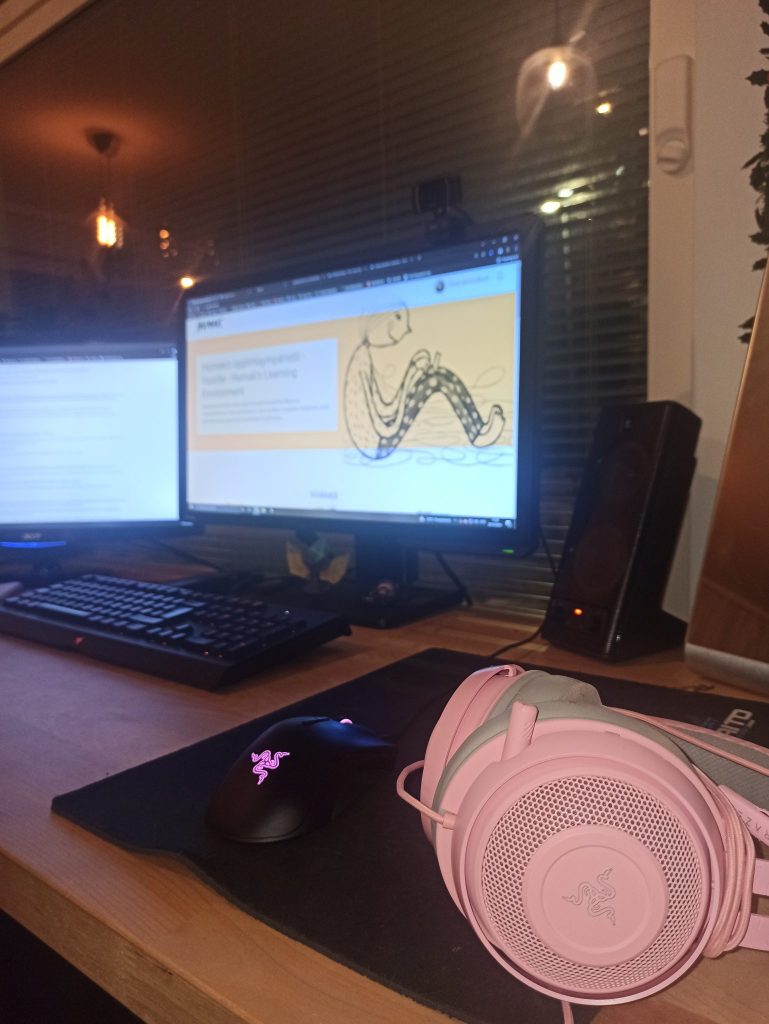 Kuvan etualalla vaaleanpunaiset kuulokkeet ja musta hiiri. Taka-alalla tietokone, jonka ruudulla näkyy Humakin verkkopohjainen oppimisympäristö.