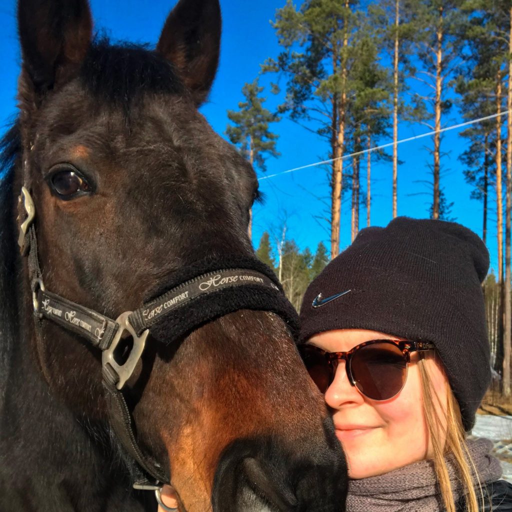 Selfie naisesta mustassa pipossa ja aurinkolaseissa halaamassa tummanruskean hevosen turpaa aurinkoista metsämaisemaa ja sinitaivasta vasten.