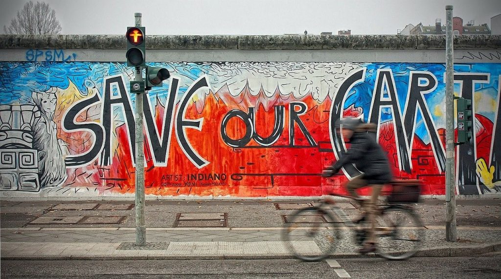 Taideteos Berliinin muurissa, jossa lukee "Safe the Earth". Muurin edessä näkyy pyöräilijän varjo.