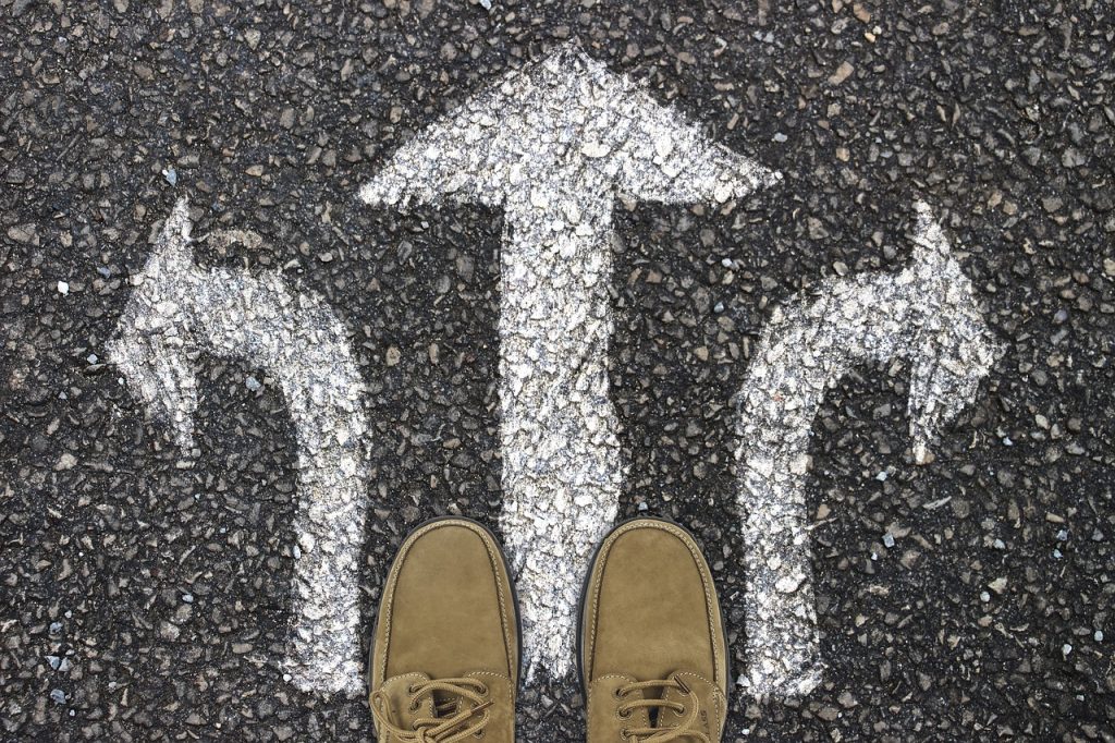 Ruskeat kengät seisovat asfaltti-tien päällä, johon on maalattu valkoisella kolme nuolta suoraan, vasemmalle ja oikealle.
