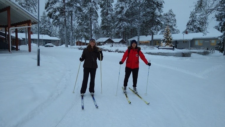 Lumisessa pihassa kaksi naista seisoo ulkovaatteissaan suksien päällä ja katsoo kameraa, kädessään heillä on sauvat. Taustalla lumisia havupuita ja taloja. 