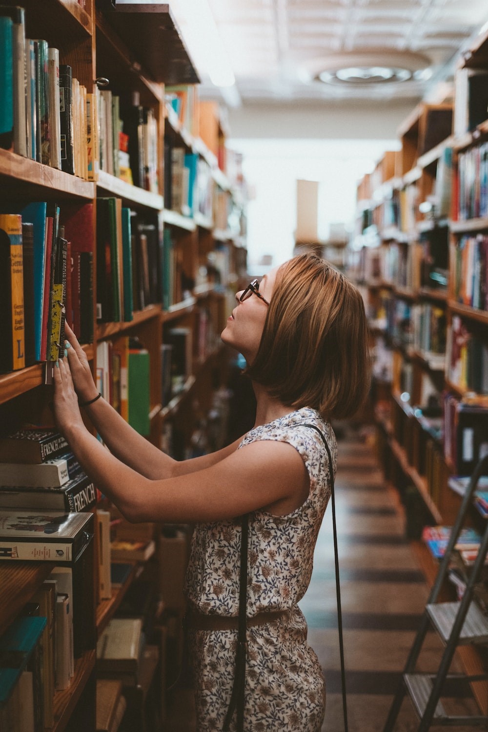 Nainen seisoo kirjastossa kirjoja täynnä olevien hyllyjen välissä ja etsii hyllyltä jotain, katse käännettynä ylöspäin, kuvakulma sivusta, kädet nojaavat hyllyn reunaan, naisella silmälasit päässään.