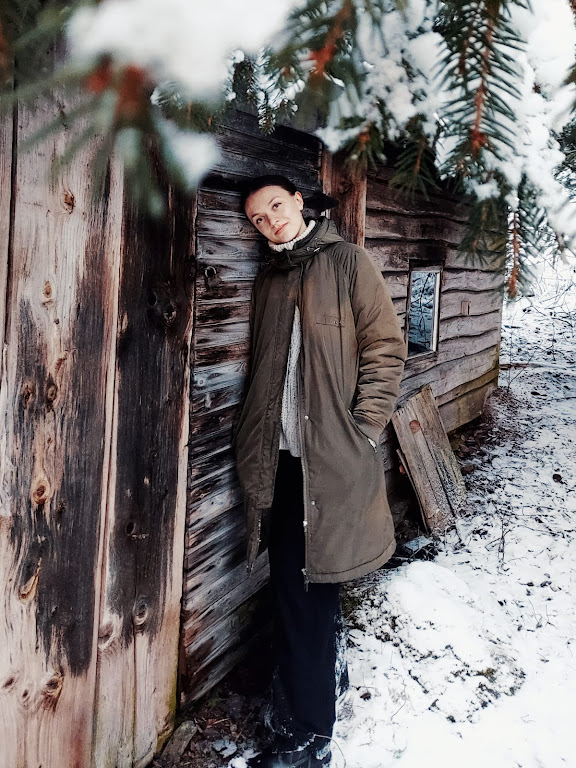 Nainen ruskeassa takissa nojaa hirsimökin oveen lumisissa maisemissa. Kuvan yläosassa pilkistää kuusen oksa.