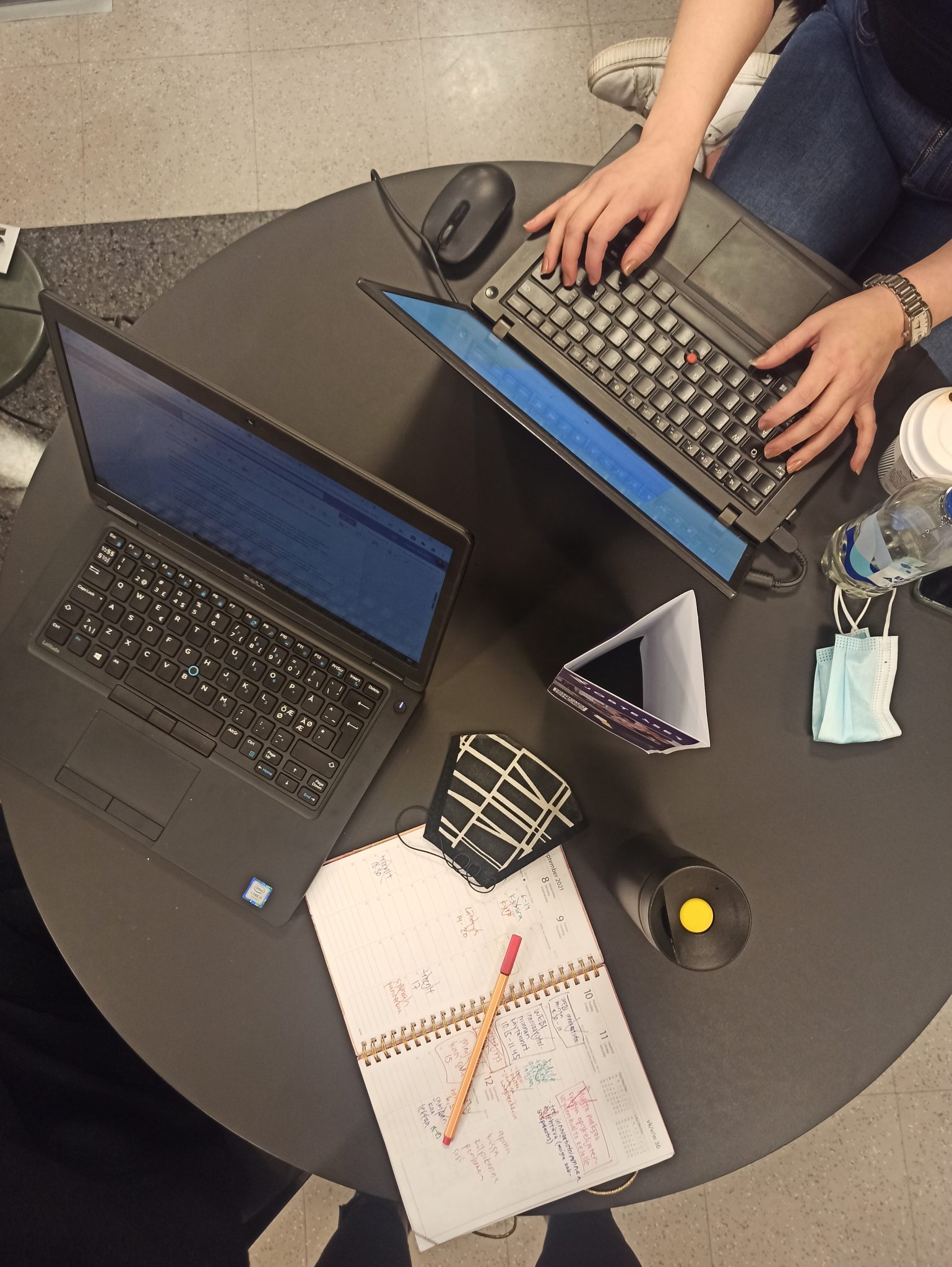 Yläperspektiivistä otettu kuva mustasta pyöreästä pöydästä, jonka päällä on kaksi tietokonetta ja opiskelutarvikkeita.