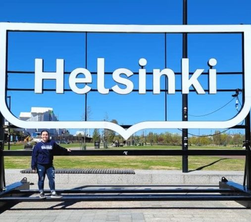 A large, white Helsinki sign pictured from the front. There is a park in the background and a dark-haired woman is standing beneath it with jeans and a hoodie on.