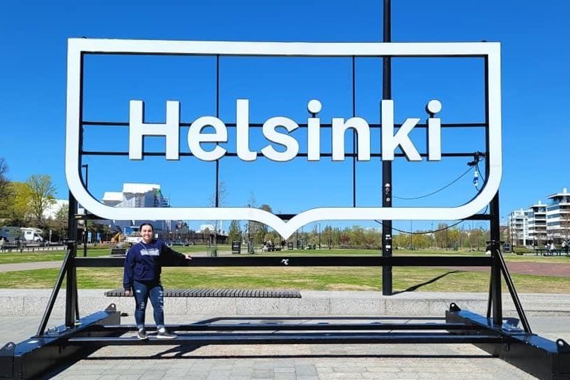 A large, white Helsinki sign pictured from the front. There is a park in the background and a dark-haired woman is standing beneath it with jeans and a hoodie on.